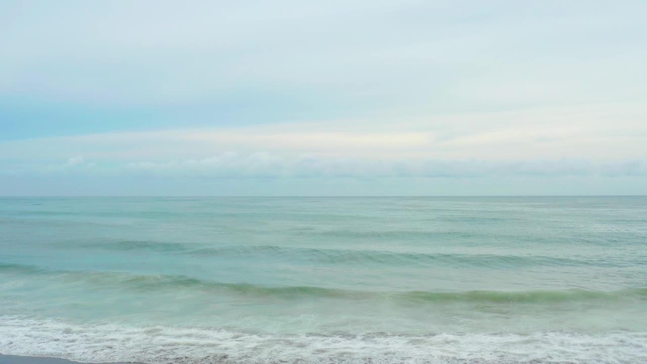 At La Playita Los Cabos, turquoise water rolls toward a foamy lower edge beneath a pale overcast sky, with no visible people.