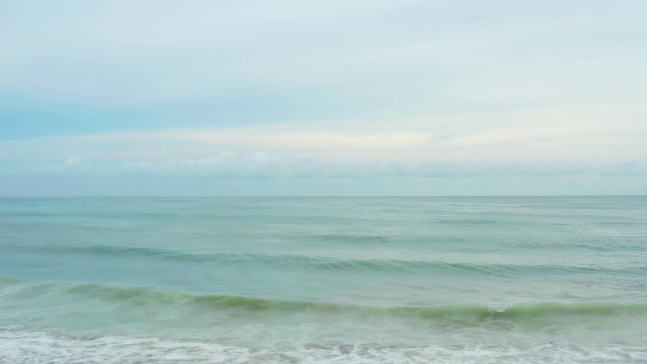 At La Playita Los Cabos, gentle wave bands stretch across open turquoise water below a soft cloud layer, with no visible people.