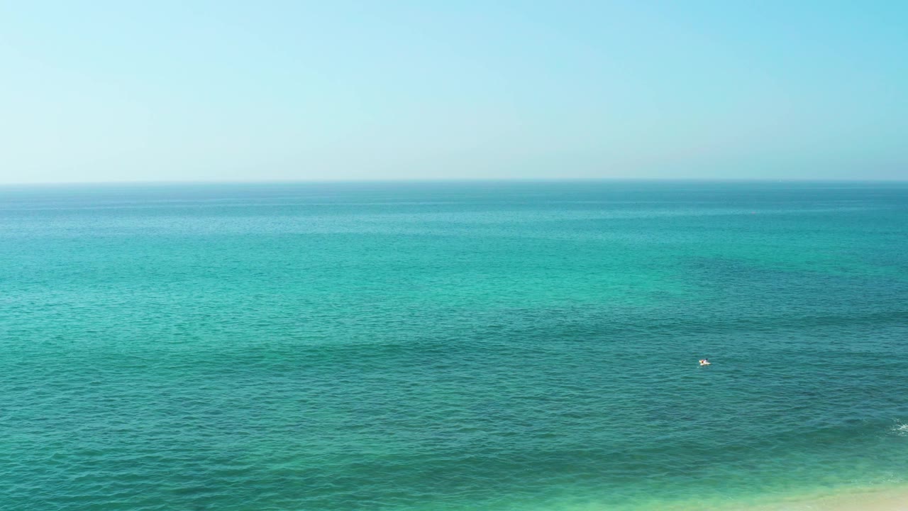 Wide view of turquoise ocean at El Tule Beach Los Cabos with a pale blue sky, faint horizon, and a tiny distant figure or object on the water.