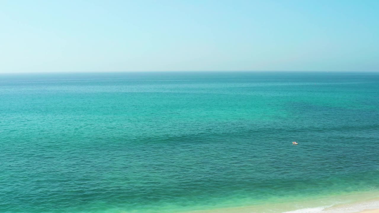 Blue-green ocean at El Tule Beach Los Cabos stretches to a soft horizon, with darker water patches and a tiny distant figure or object on the surface.