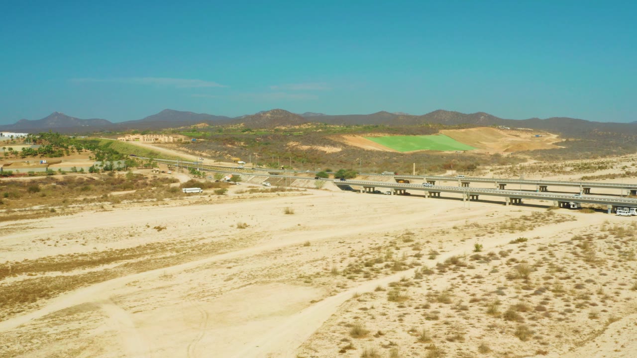 At one second, the El Tule Beach Los Cabos aerial view holds the overpass low in frame, with pale sand, scrub patches, brown hills, a green field, and no visible people.