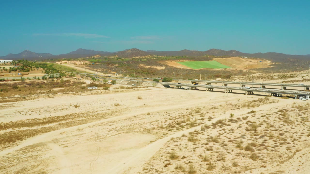 At three seconds, the El Tule Beach Los Cabos frame emphasizes the empty sandy foreground and low scrub, with the overpass and small white vehicle set below distant hills and a clear blue sky, with no visible people.