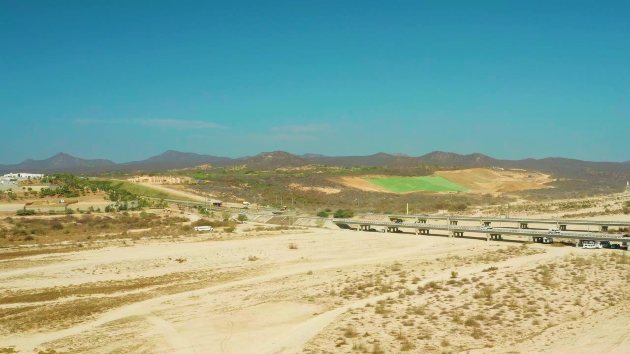 Aerial frame at El Tule Beach Los Cabos with the bridge stretched across the sandy wash, vehicles on the roadway, pale hills, dark mountain ridges, and no visible people.