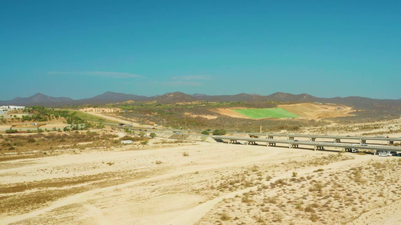 Wide aerial frame from El Tule Beach Los Cabos with the dry wash in front, bridge and vehicles across the lower midground, tan hills, dark mountains, and no visible people.