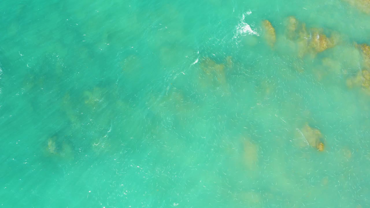Overhead view of turquoise water at El Tule Beach Los Cabos with faint submerged rocks near the upper frame and no visible people.