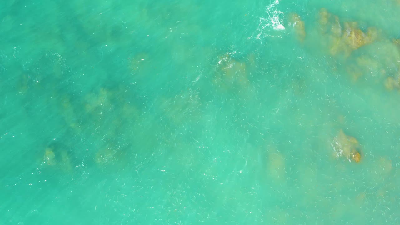 Top-down turquoise sea at El Tule Beach Los Cabos with pale and darker underwater areas, scattered rocks, and no visible people.