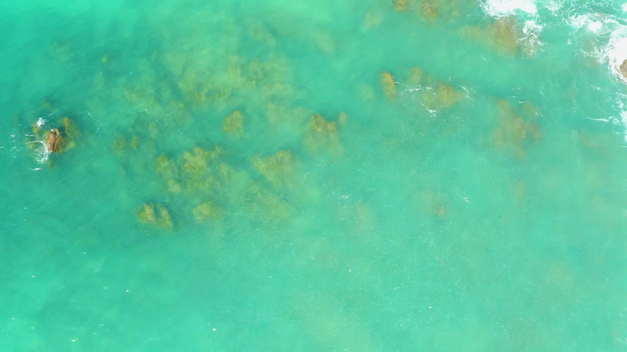 Clear blue-green ocean water at El Tule Beach Los Cabos shows scattered submerged rocks beneath a lightly rippled surface, with no visible people.
