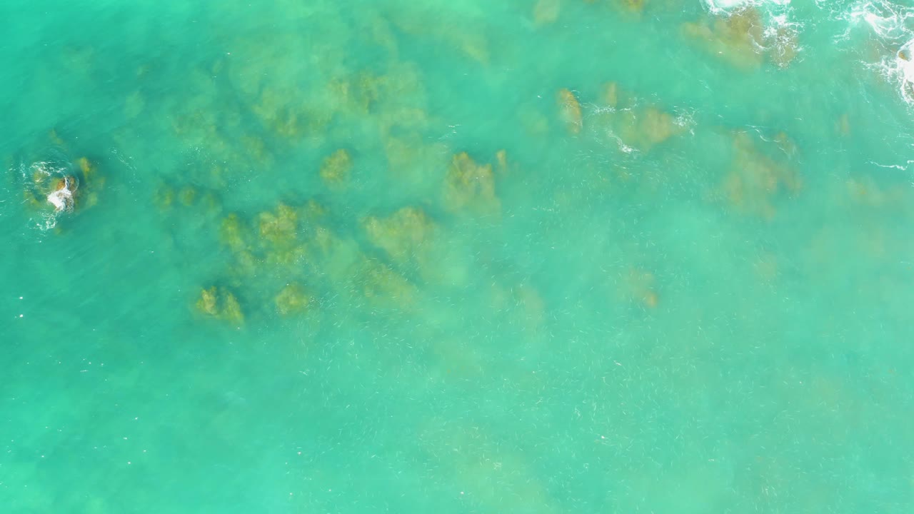 Aerial-looking view of shallow turquoise water at El Tule Beach Los Cabos with darker and tan rock patches visible below the surface, with no visible people.