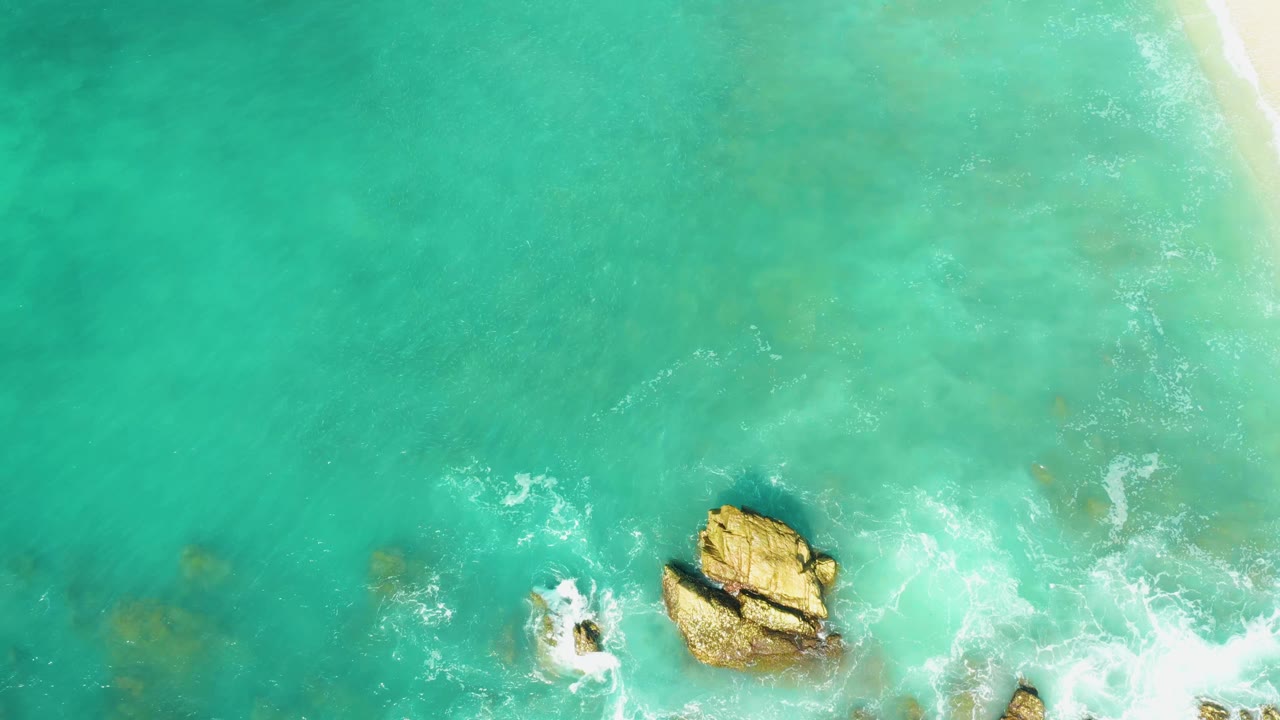 At 0:01, El Tule Beach water fills the frame with the largest rock low center, a smaller rock to the left, white surf curling around both, and no visible people.