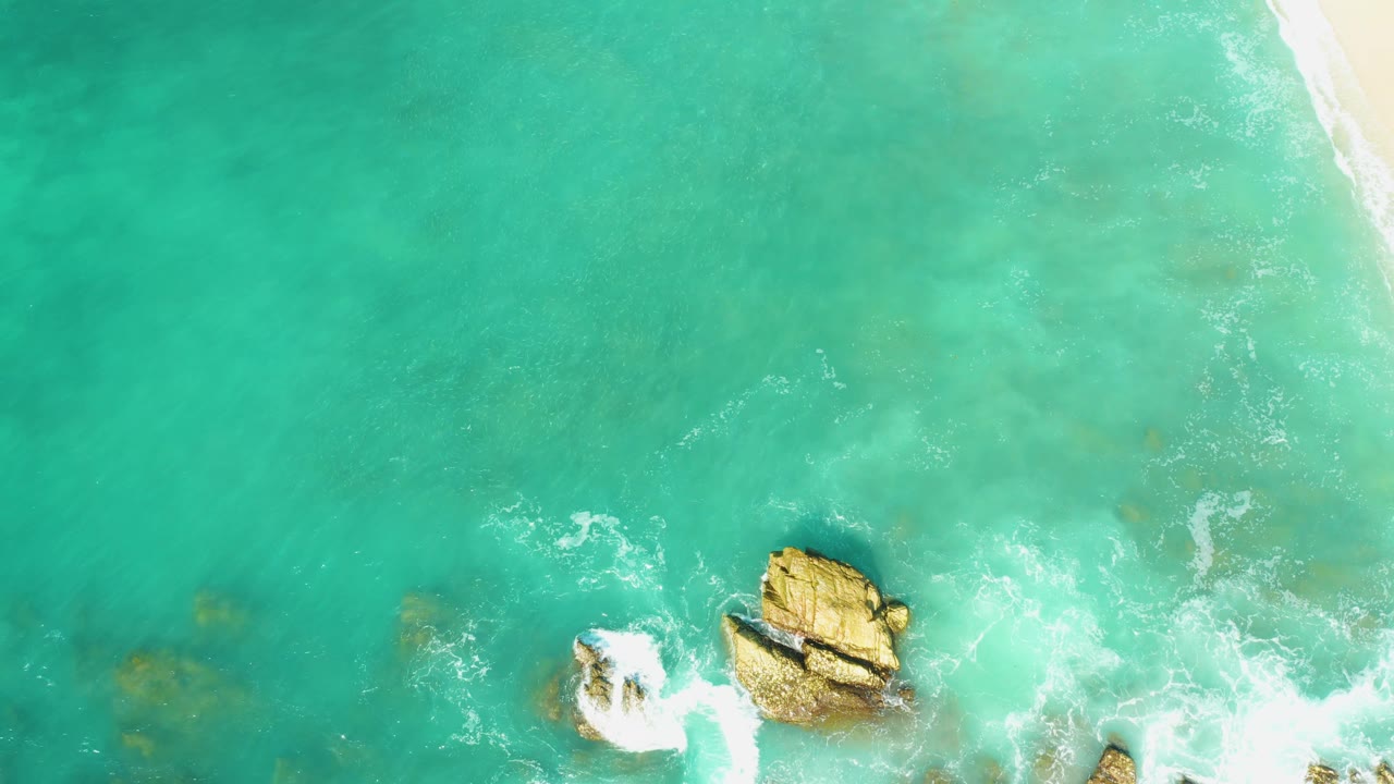 At 0:02, the overhead view shows green-blue water, two rocky outcrops near the lower half, a thin sandy shoreline along the right edge, and no visible people.