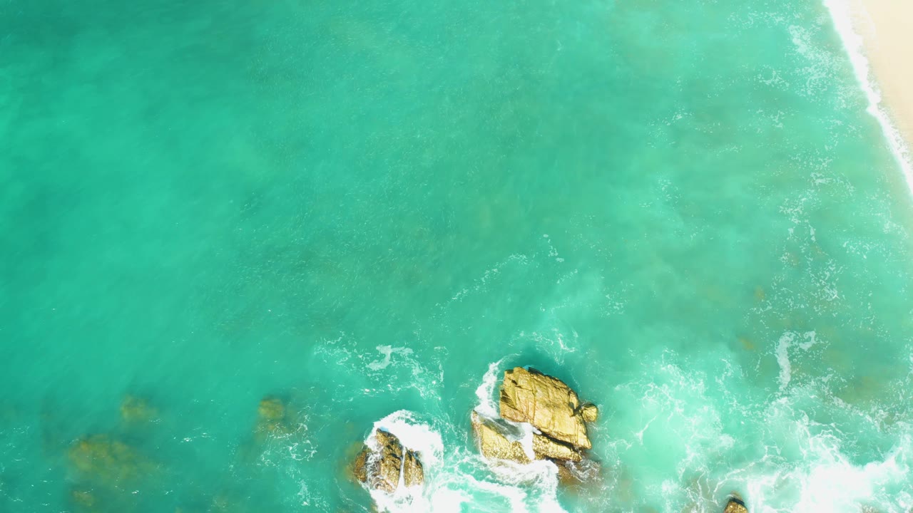 At 0:03, El Tule Beach appears in a top-down composition with the main rock closer to the lower edge, foamy water surrounding it, pale sand at right, and no visible people.