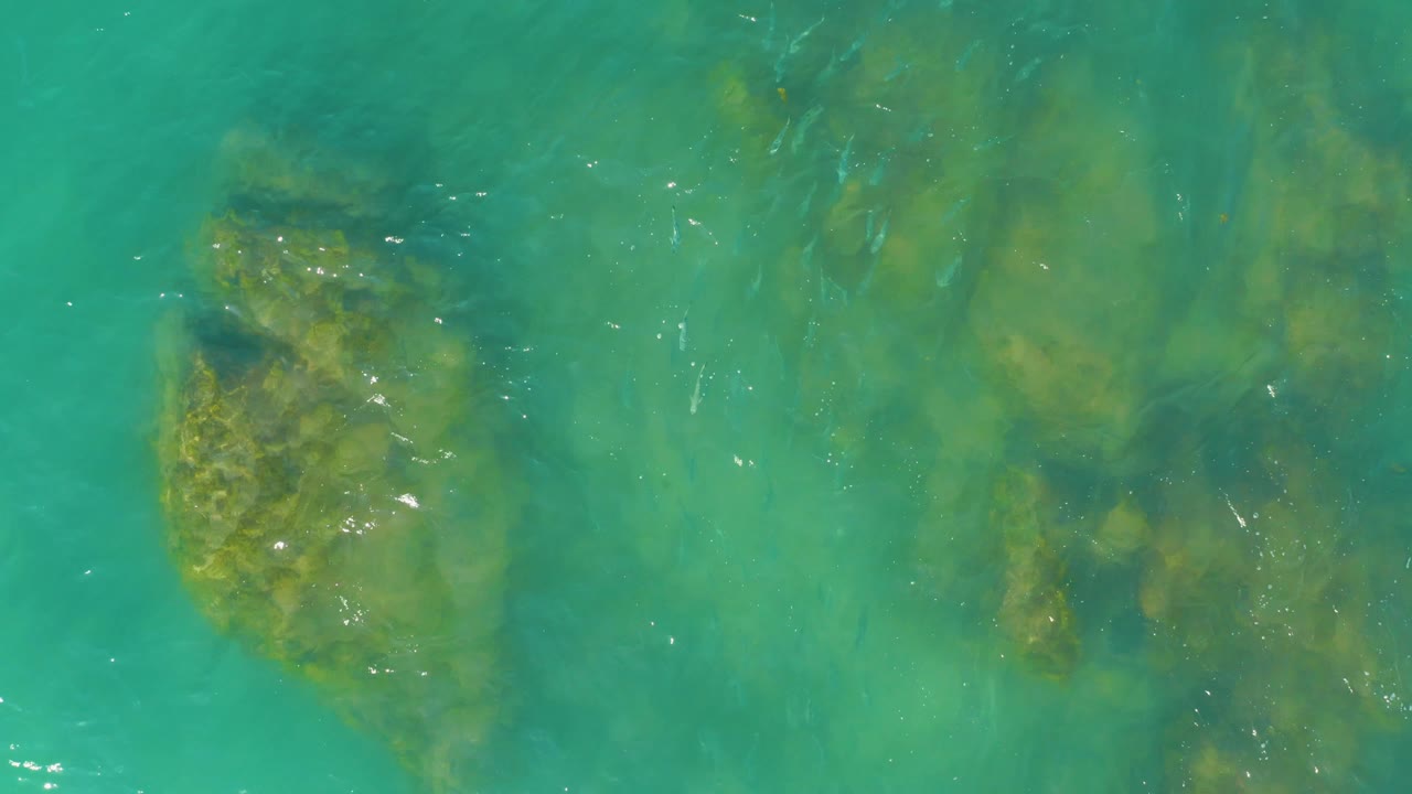 One second in, clear turquoise water at El Tule Beach shows a large green rock shape on the left and softer submerged forms to the right, with no visible people.