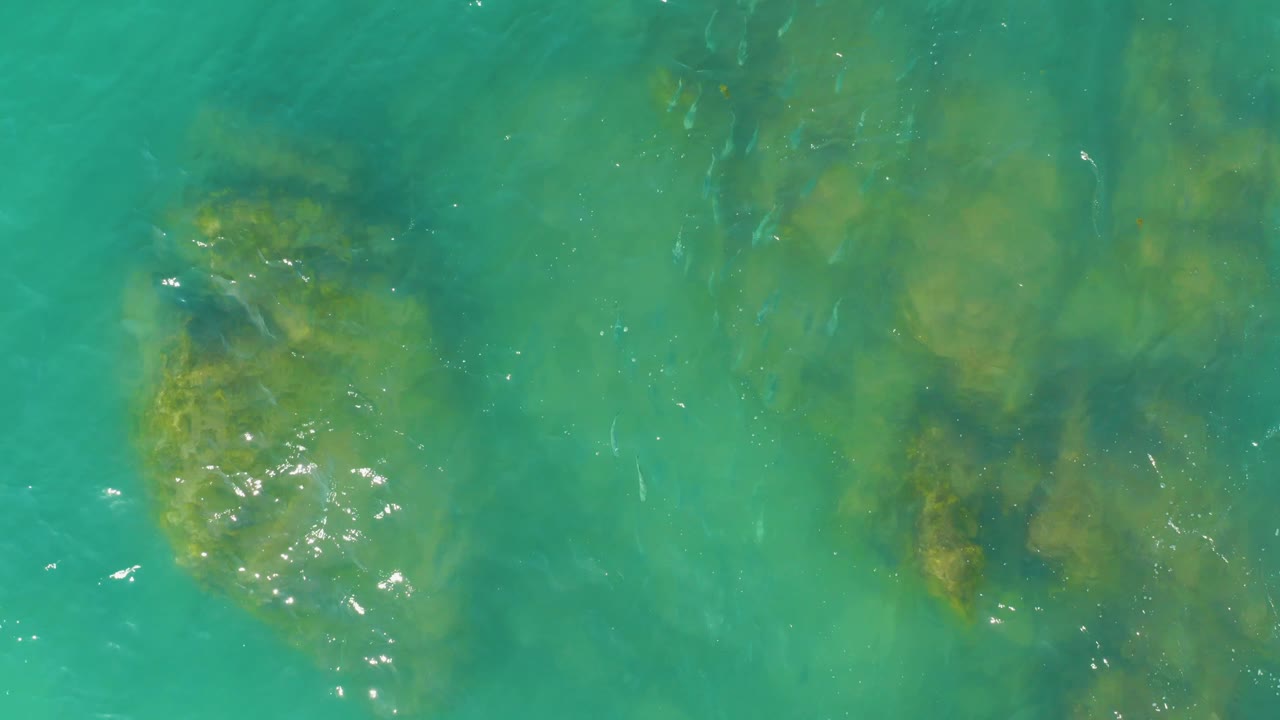 At 0:02, the left submerged rock appears brighter under the rippled turquoise surface while darker green patches sit deeper to the right, with no visible people.