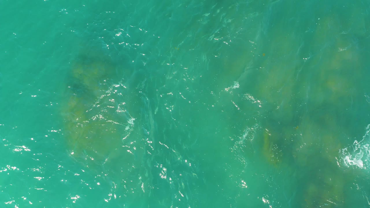 Overhead view of turquoise water at El Tule Beach in Los Cabos, with faint yellow-green submerged rock patches under small surface ripples and no visible people.