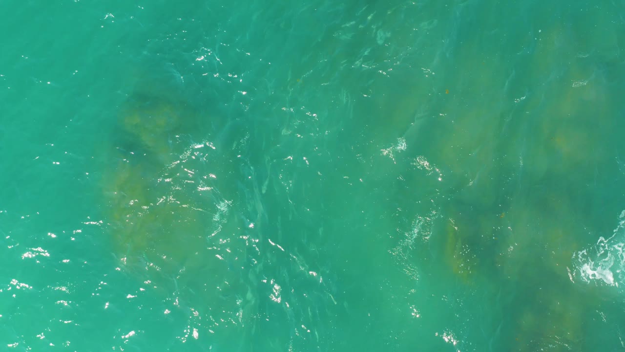 Blue-green water fills the frame at El Tule Beach in Los Cabos, where submerged rock patches appear through shifting ripples and small white highlights, with no visible people.