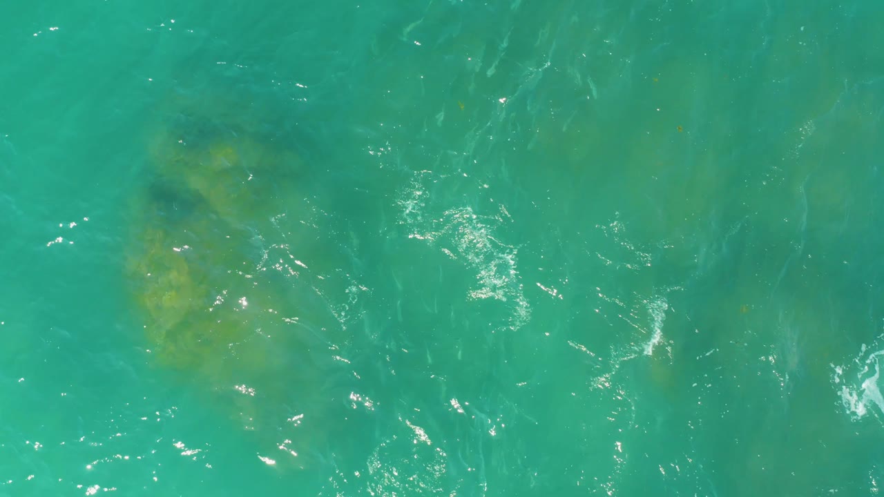 A larger yellow-green submerged formation sits near the upper right beneath turquoise water at El Tule Beach in Los Cabos, with ripples and no visible people.