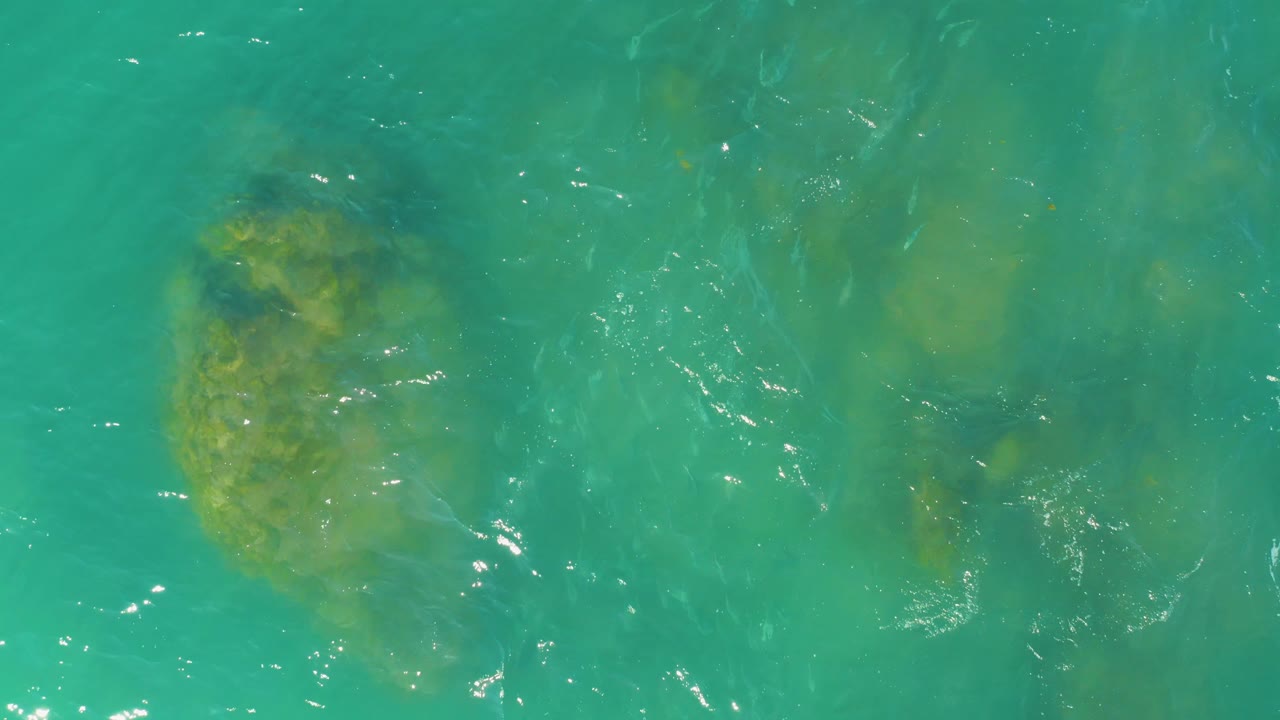 Teal ocean water at El Tule Beach in Los Cabos reveals scattered submerged rocks below soft surface movement, with a brighter rock patch at the left and no visible people.