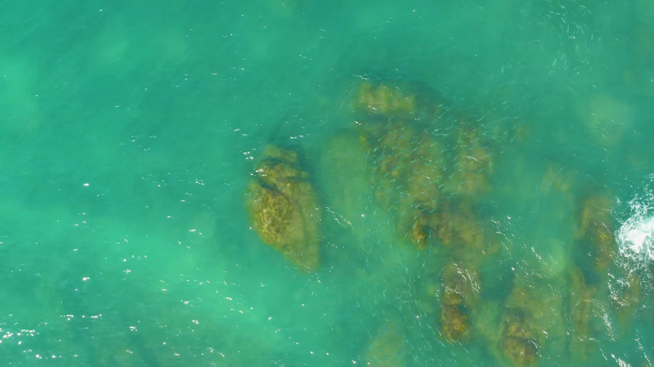 Aerial overhead view at El Tule Beach, Los Cabos, showing clustered submerged rocks under green-blue water, moving surface streaks, white foam at the right edge, and no visible people.