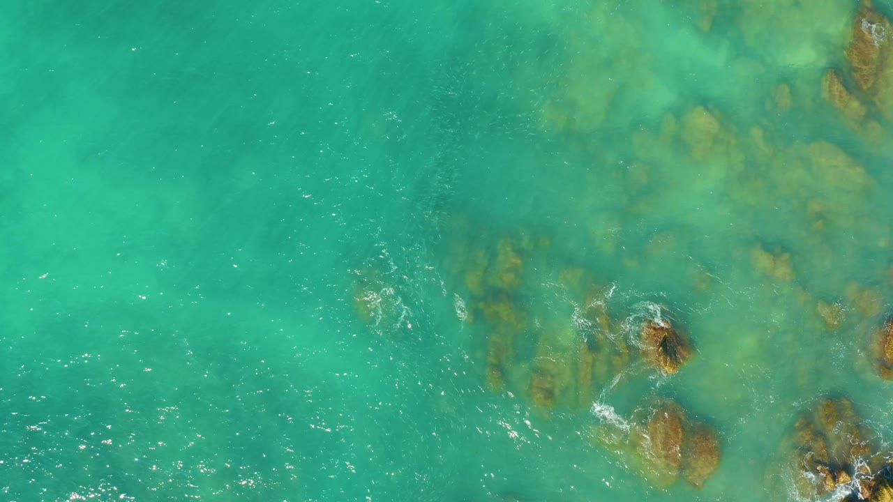 Overhead view of turquoise water at El Tule Beach Los Cabos flowing over brown submerged rocks, with small foam patches and no visible people.
