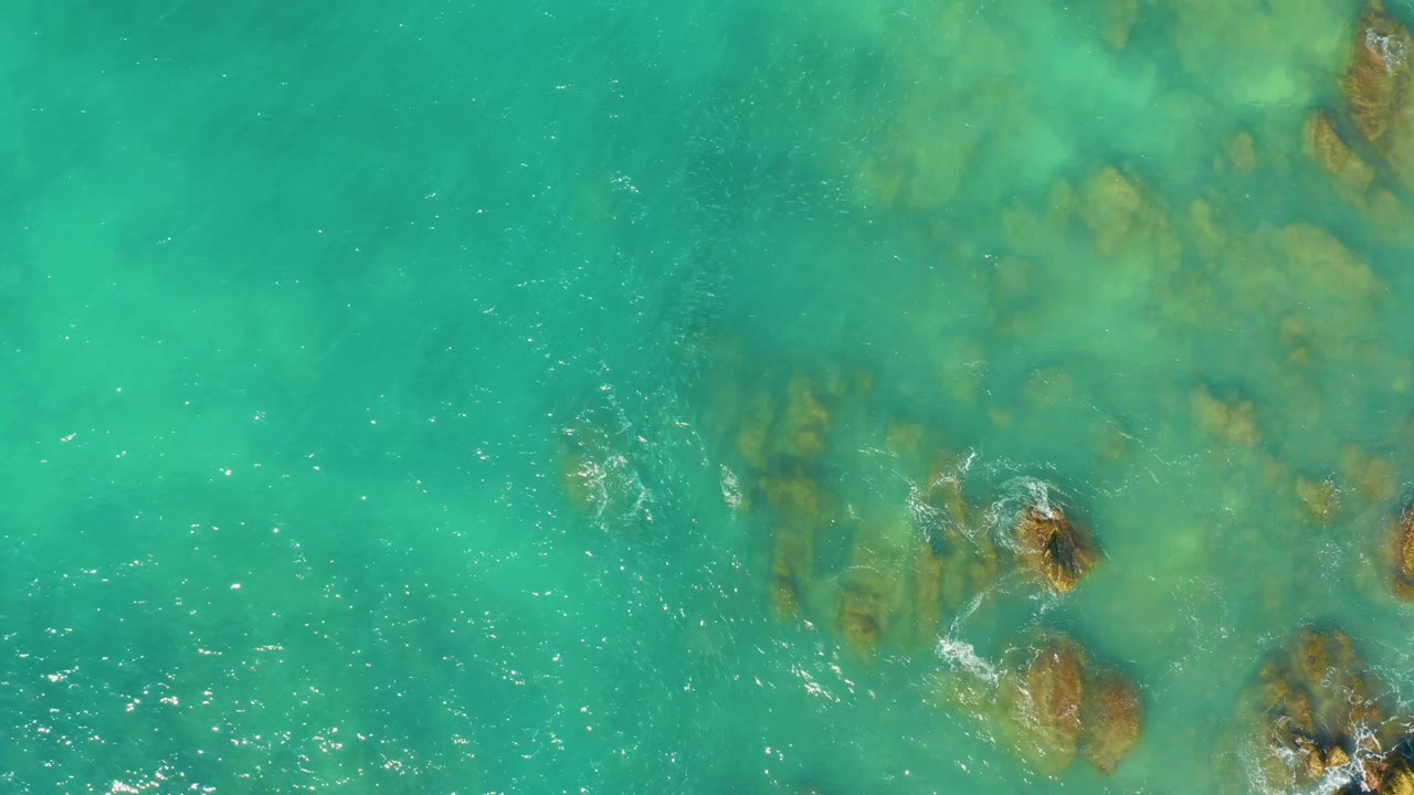 Clear turquoise sea at El Tule Beach Los Cabos shows submerged rock shapes beneath the surface, with scattered white foam drifting across the left side and no visible people.