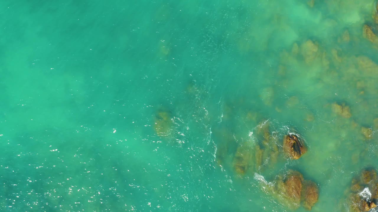 Top-down frame at El Tule Beach Los Cabos with turquoise water covering clustered brown rocks, light foam trails near the right edge, and no visible people.