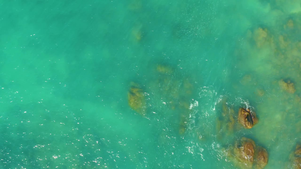 Green-blue water at El Tule Beach Los Cabos covers several submerged rocks, while a small white foam break forms near a darker rock and no visible people appear.