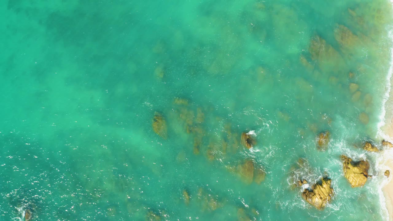 Aerial view over El Tule Beach turquoise water, with submerged tan rocks, small white foam breaks, a sandy shoreline strip at right, and no visible people.