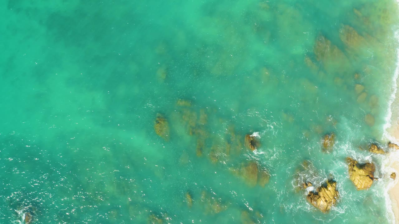 Clear turquoise sea with underwater rocks and white foam around shoreline rocks.