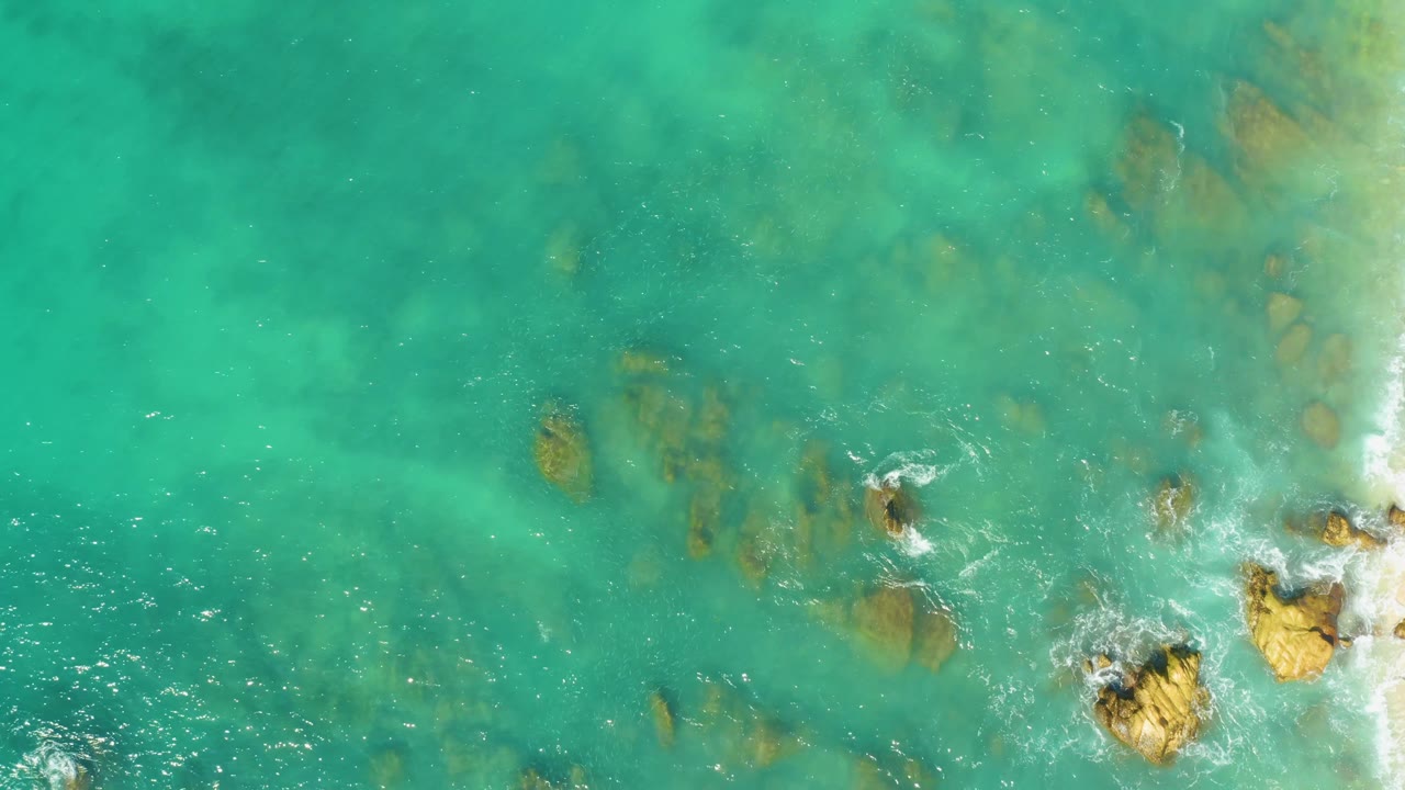 Aerial frame from El Tule Beach showing turquoise ocean shallows, brown rock shapes under the surface, active foam at the right edge, and no visible people.