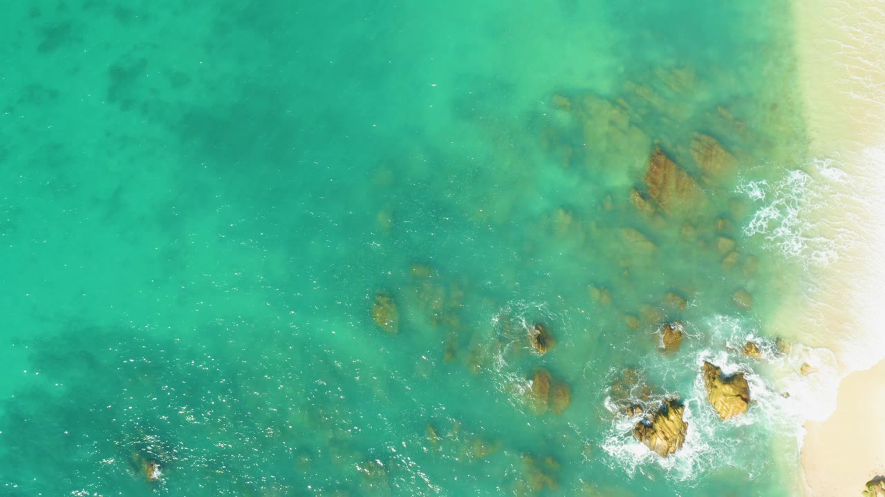Aerial frame at El Tule Beach Los Cabos showing clear green-blue water, rock clusters near the shore, small white surf patches, sand along the right edge, and no visible people.