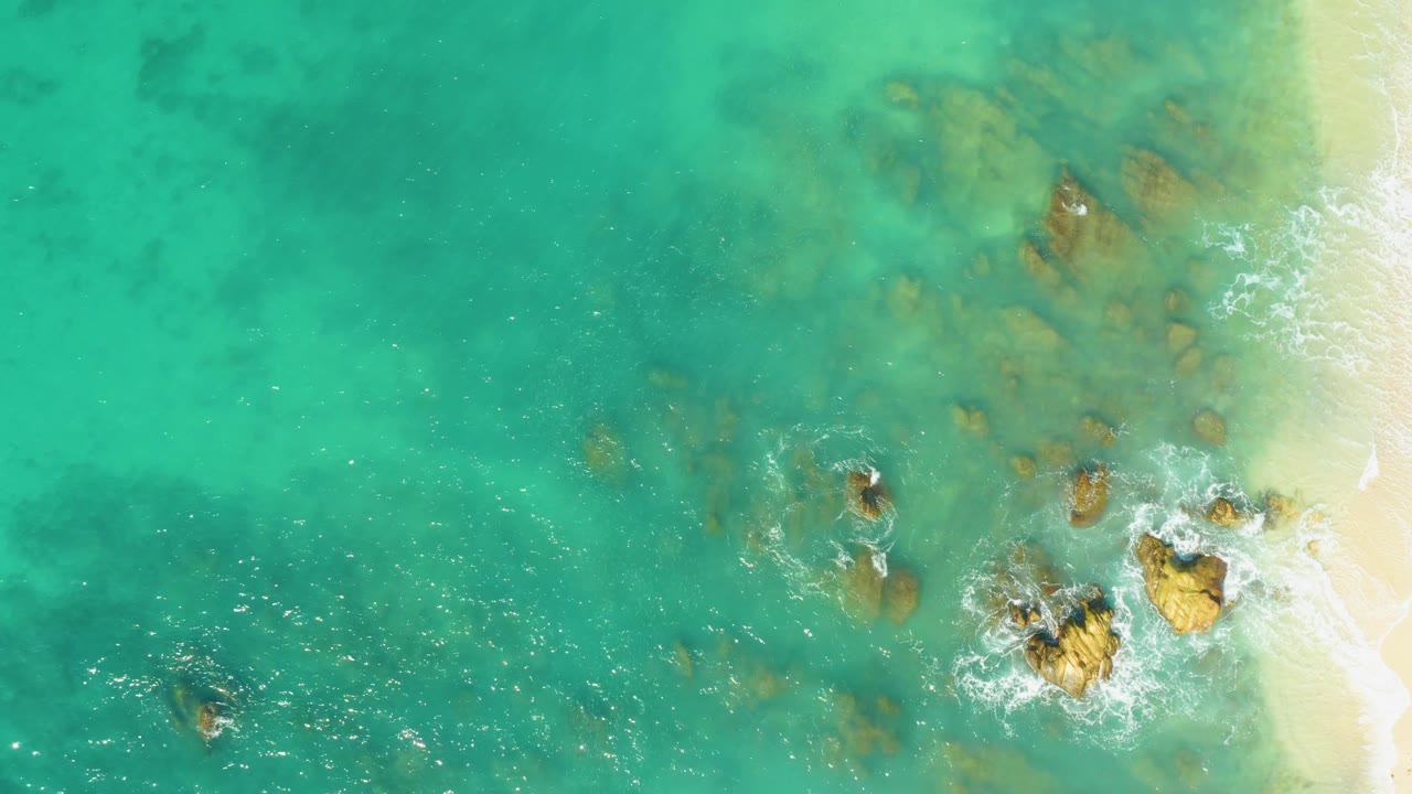 El Tule Beach Los Cabos appears from above with shallow turquoise water, softened rock shapes below the surface, a narrow sand strip on the right, light foam at the rock edges, and no visible people.
