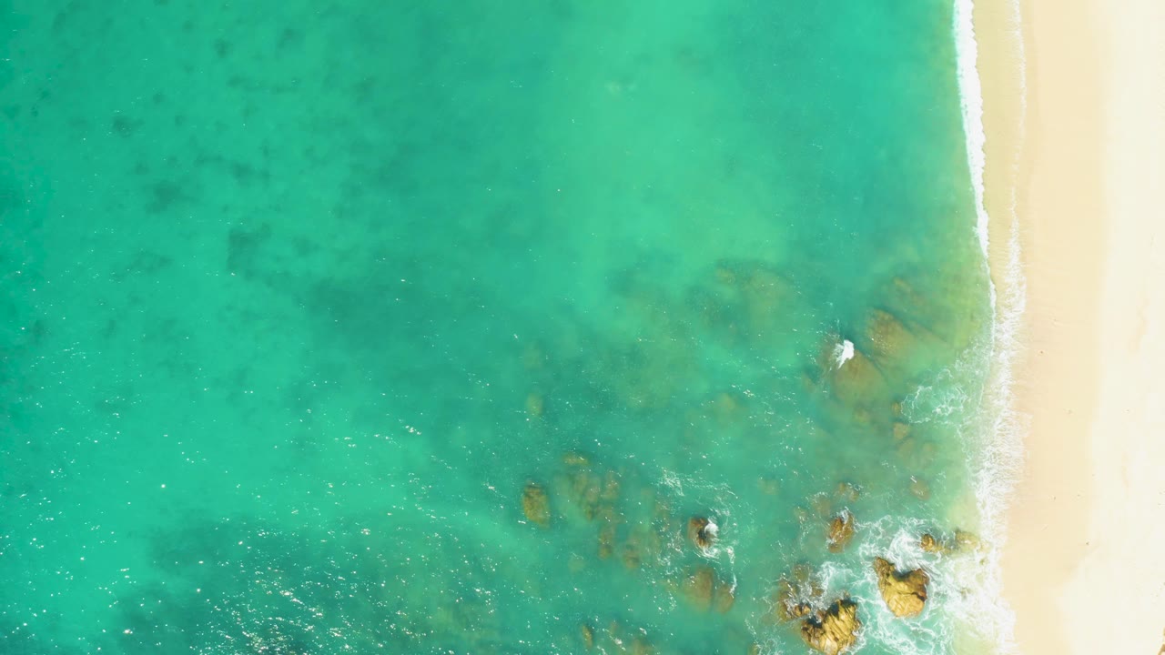 Overhead view at El Tule Beach Los Cabos showing turquoise water, pale sand along the right edge, submerged rocks near the lower center, small foam patches, and no visible people.