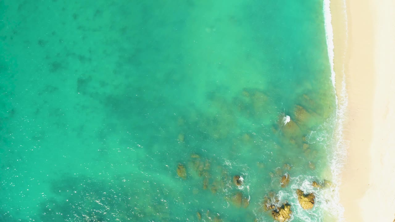 At 0:01, the frame holds on clear green-blue water at El Tule Beach Los Cabos, with sand tight to the right side, rocks below the surface, light foam, and no visible people.