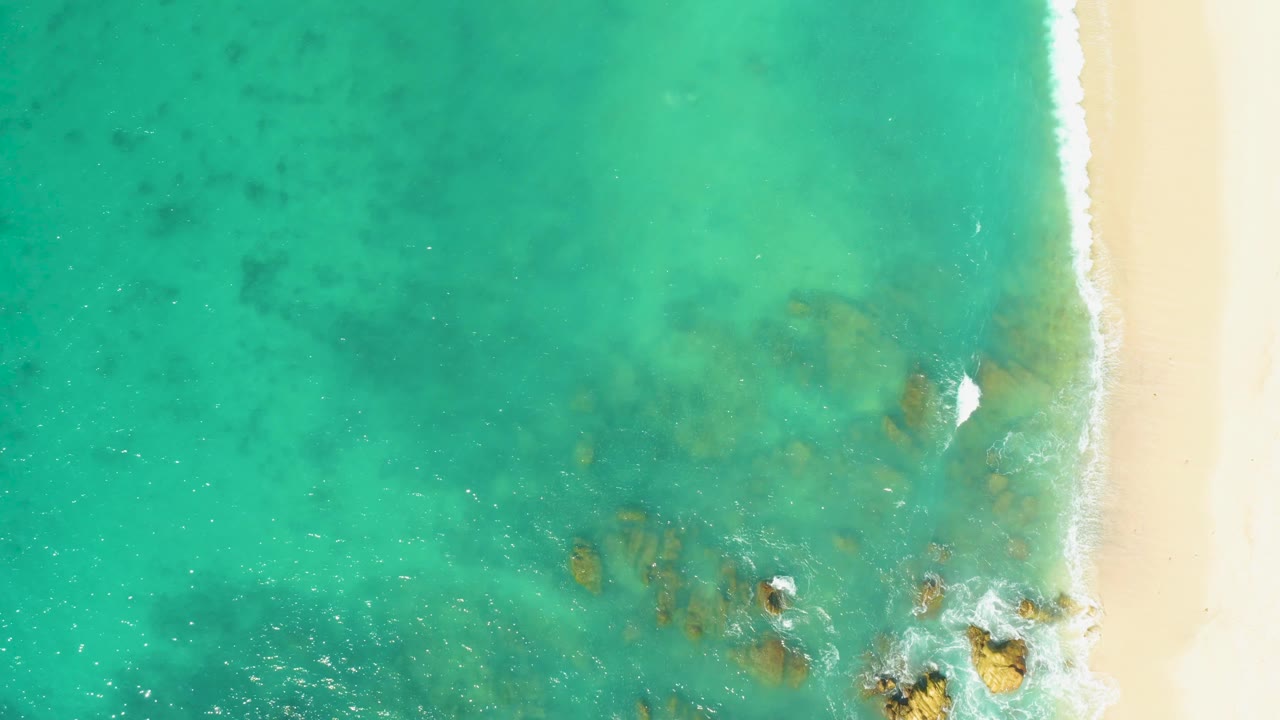 At 0:02, turquoise water fills most of the El Tule Beach Los Cabos frame while a narrow pale beach strip runs down the right, with rock clusters and white foam near the lower right, no visible people.