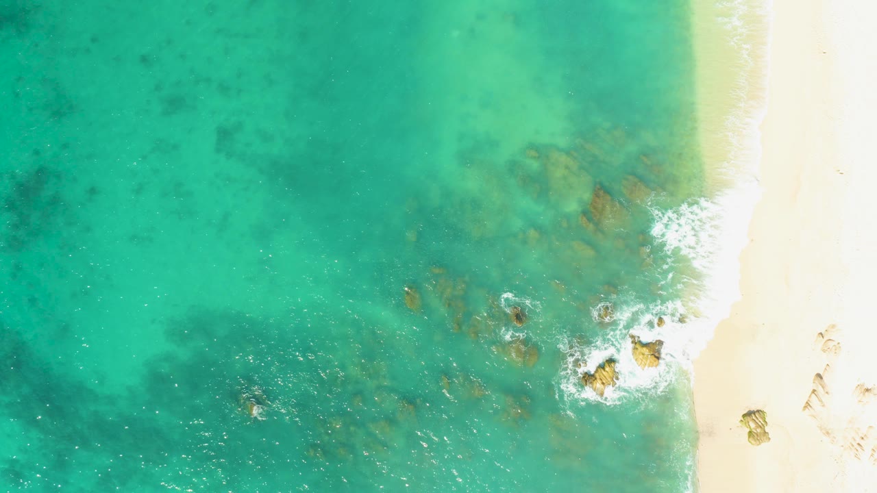 Overhead frame of El Tule Beach in Los Cabos with turquoise water, submerged rocks near the shore, white foam at the sand line, and no visible people.