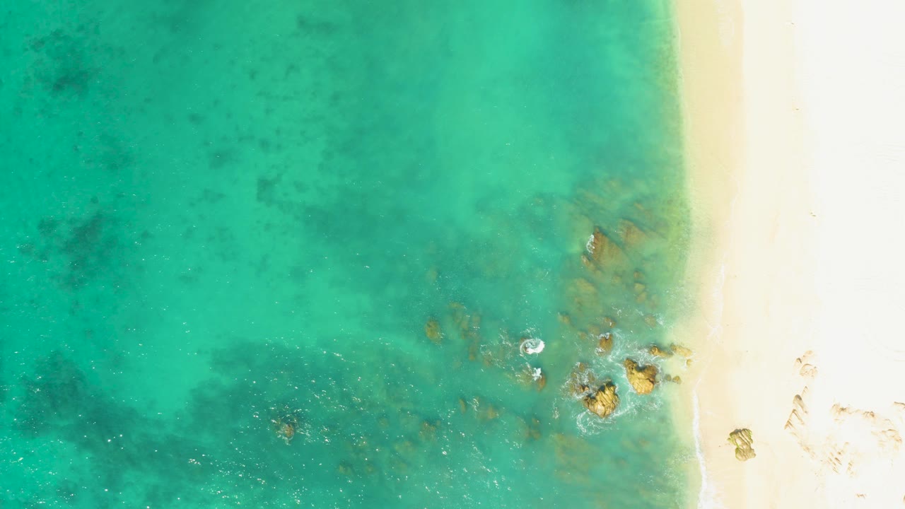 Top-down view of El Tule Beach in Los Cabos with turquoise water, pale sand along the right edge, scattered rocks near the shoreline, light foam, and no visible people.