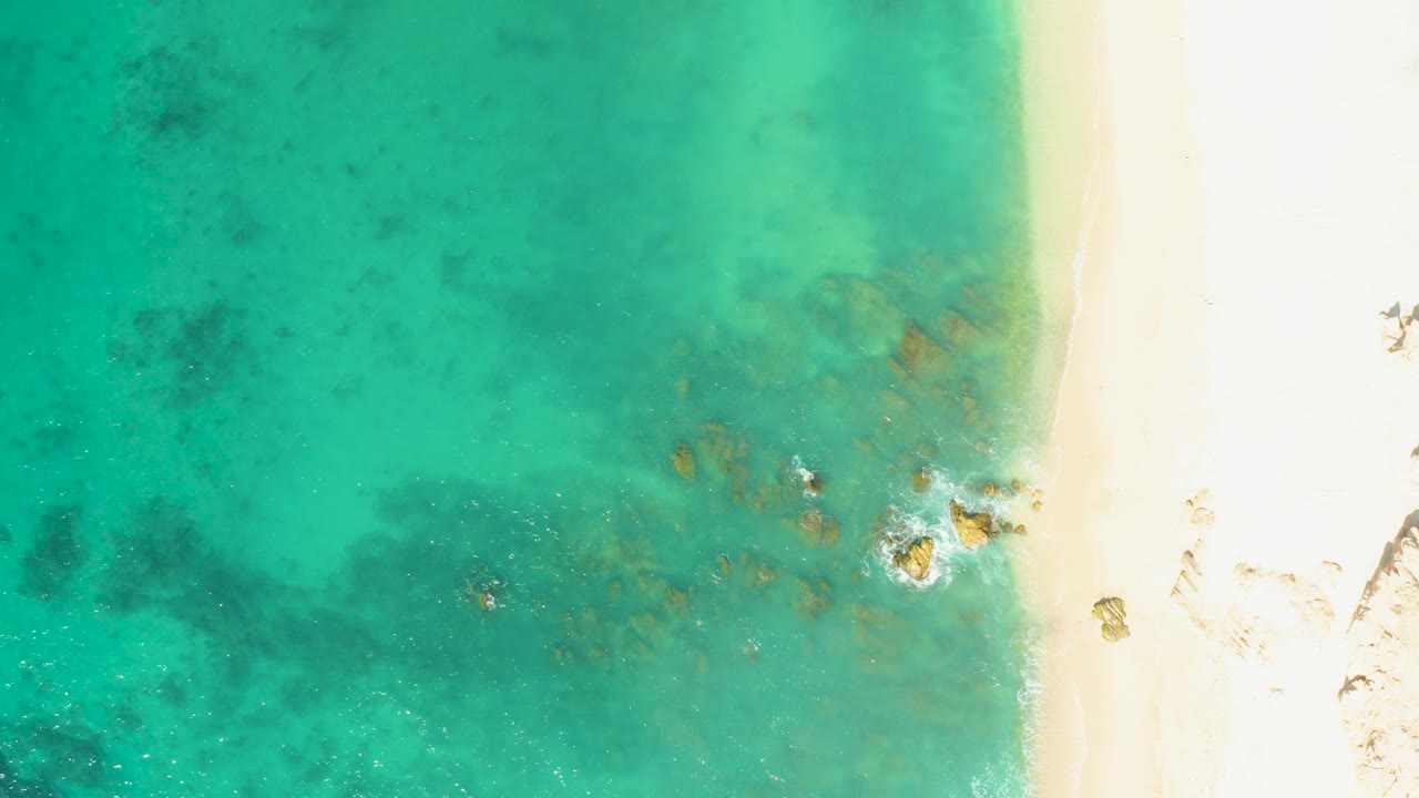 El Tule Beach shoreline seen from above, with pale sand running vertically, clear green-blue water, small foam patches around rocks, and no visible people.