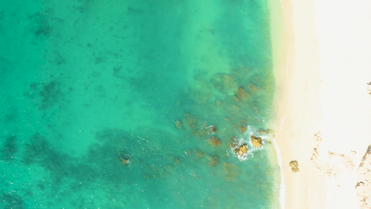Overhead El Tule Beach view with a cluster of submerged rocks near the lower right shoreline, clear water across the frame, bright sand, and no visible people.
