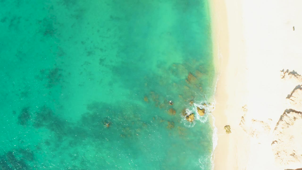 Aerial frame at El Tule Beach in Los Cabos with clear blue-green water filling the left side, bright sand along the right edge, scattered underwater dark patches, and no visible people.