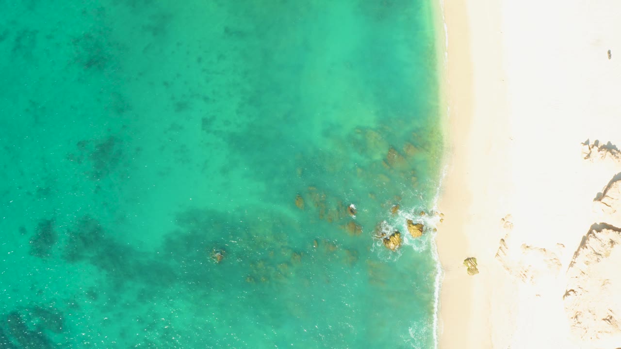 Aerial frame at El Tule Beach in Los Cabos with shallow blue-green water across the left and center, pale sand along the right, soft wave texture at the edge, and no visible people.