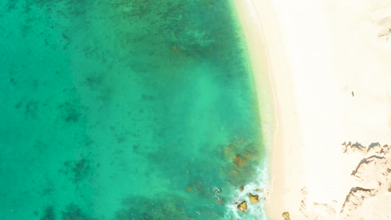 At 0:01, the overhead El Tule Beach Los Cabos view keeps the shoreline on the right while clear turquoise water and darker submerged patches fill the left side, with no visible people.