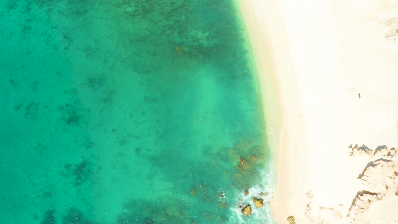 At 0:02, turquoise shallows dominate the frame, the pale beach remains along the right edge, and a small rocky surf area sits near the lower right with no visible people.