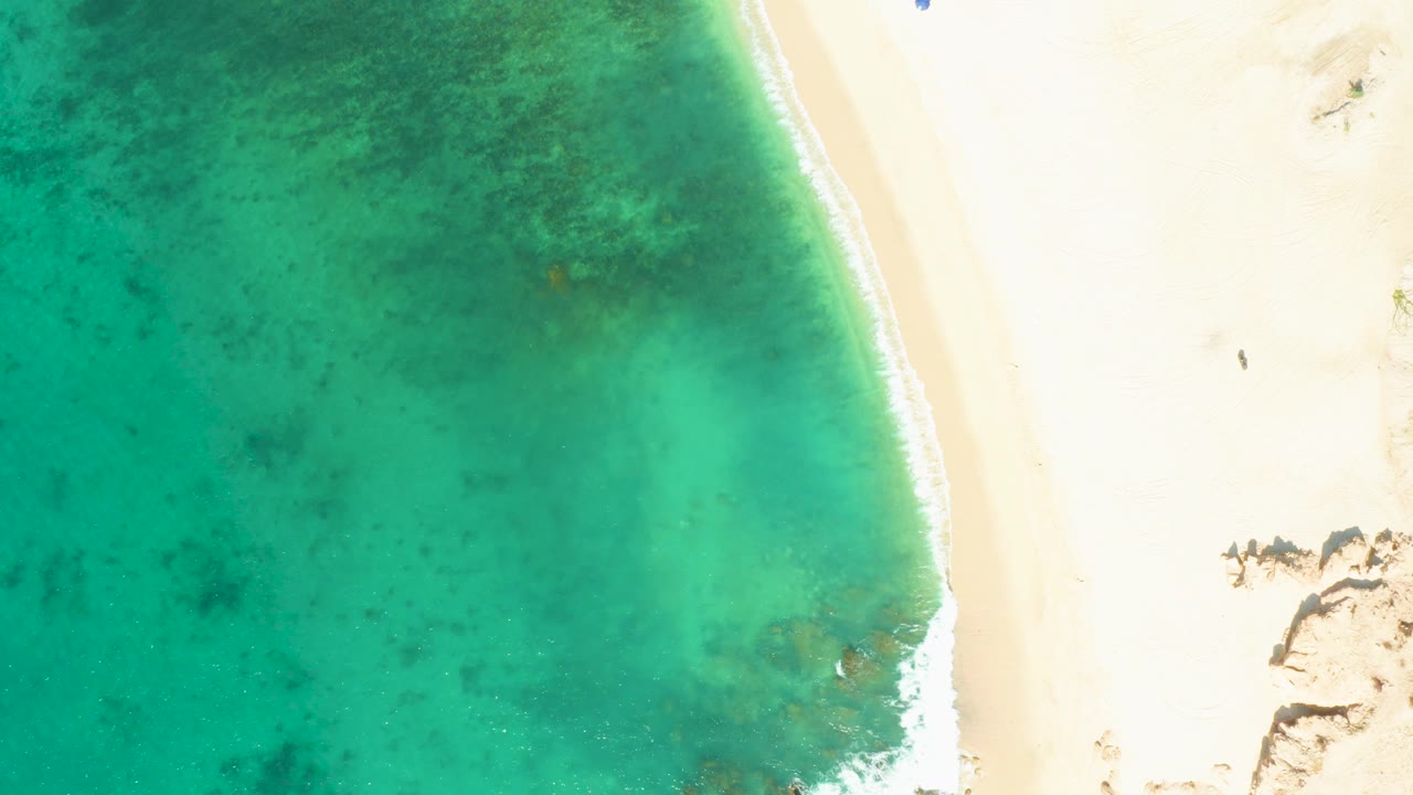 Aerial frame from El Tule Beach in Los Cabos with the shoreline curving down the right side, green-blue water filling the left, rocks near the bottom right, and no visible people.