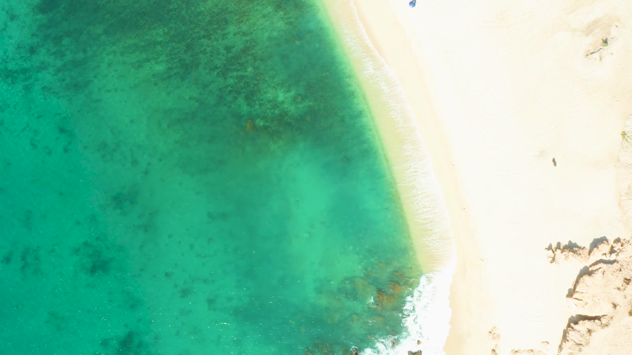 Overhead shot at El Tule Beach in Los Cabos where darker turquoise water spreads across the left, the sandy shore runs along the right, rocks sit near the lower right, and no visible people appear.
