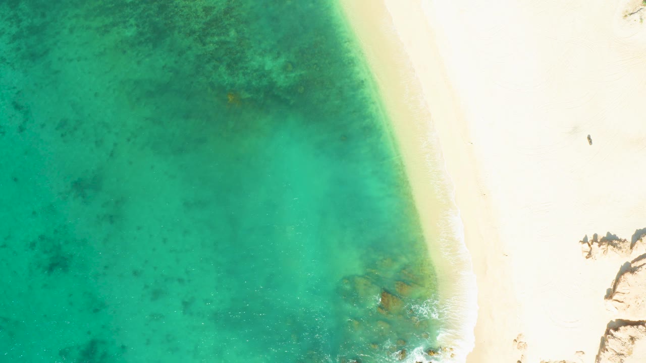 At 0:01, El Tule Beach appears from above with the surf line curving along pale sand and darker underwater texture visible through the turquoise water, with no visible people.