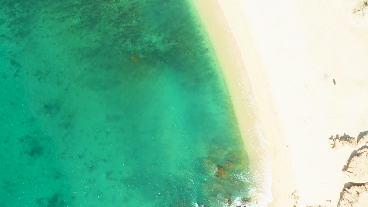 At 0:02, the overhead view shows turquoise water pressing toward the beach, a pale sand strip, small foam detail, and no visible people on the El Tule Beach shoreline.