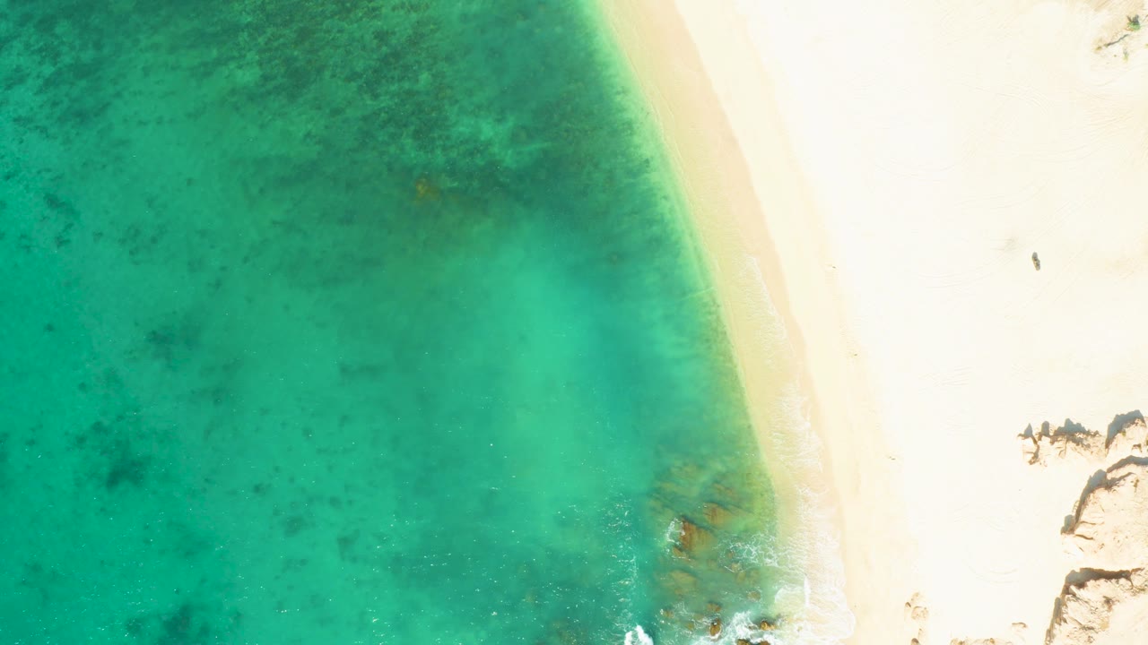 At 0:03, shallow turquoise water fills most of the left side while empty bright sand and a thin wave edge sit to the right at El Tule Beach, with no visible people.
