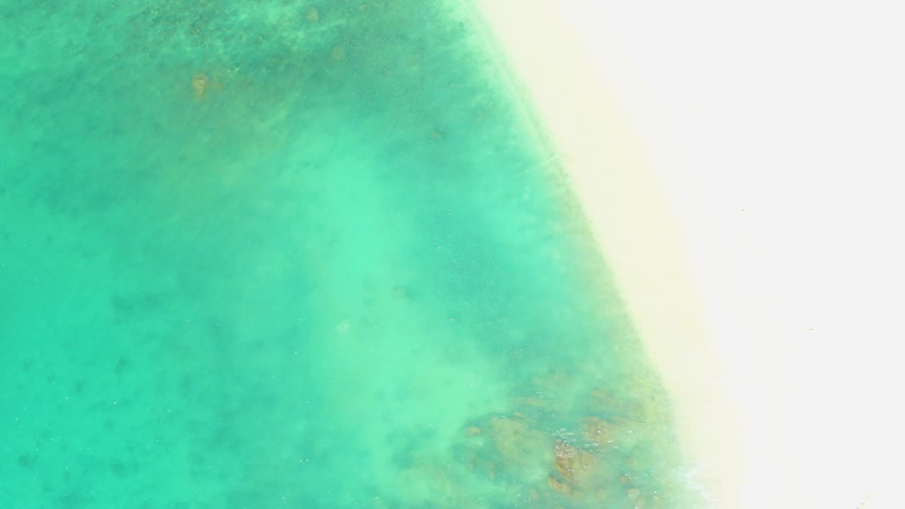 At 0:01, the aerial view keeps the pale sand edge on the right while clear green-blue water and faint seabed texture fill most of the frame, with no visible people.