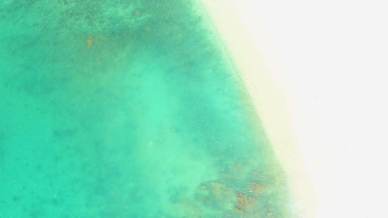 At 0:02, shallow turquoise water covers the left side of the El Tule Beach frame, with the bright sandy shore at right and small darker patches under the surface, with no visible people.