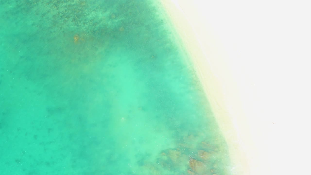 At 0:03, the top-down view shows the sandy beach band narrowing along the right edge as turquoise water and muted seabed marks occupy the frame, with no visible people.
